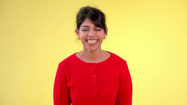 A woman in a red cardigan making a shushing gesture against a bright yellow background. She has dark hair and is smiling playfully, conveying a sense of secrecy or playfulness.