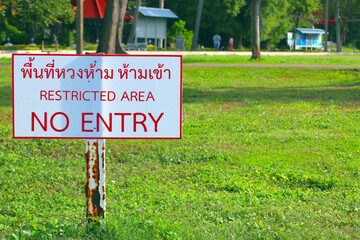 A bilingual “Do Not Enter” sign sits on a pole in the rusty grass area. The Thai-English warning sign is clearly visible. There is a park-like area and buildings behind it.