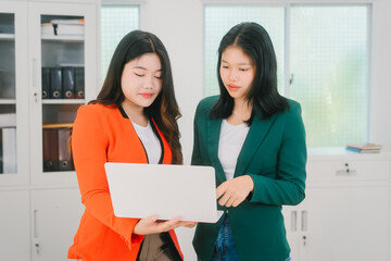 Two Asian business woman are discussing business reports and analyzing data at a desk. One is using...
