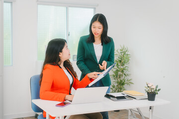 Two Asian business woman are discussing business reports and analyzing data at a desk. One is using...