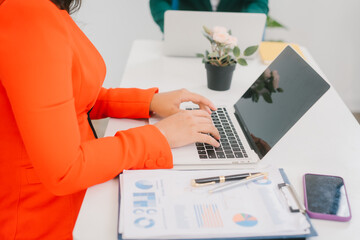 Two Asian business woman are discussing business reports and analyzing data at a desk. One is using a tablet while the other gestures. They appear focused,exchanging ideas in a modern office setting