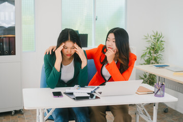 A stressed businesswoman holds her head in frustration while her colleague offers support by placing a comforting hand on her shoulder. They are working in an office with laptops and tablets