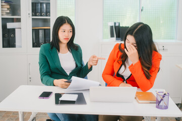 Two Asian women sit at a work desk in a tense atmosphere. They hold their heads and show signs of worry or exhaustion, with documents and phones indicating a difficult work moment