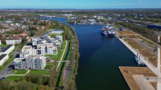 Drone flying over Canal de Caen &agrave; la Mer, docked ships, modern residential buildings, and distant Viaduc de Calix bridge, industrial and urban landscape, Normandy, France. Aerial drone forward