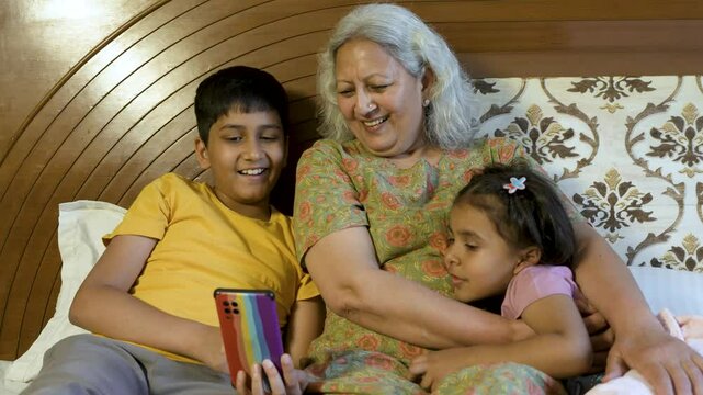 Indian grandmother with grandparents using smart phone at home. Children with grandma watching video or social media on mobile phone. 5G fast internet. Entertainment. Retired senior woman with kids.