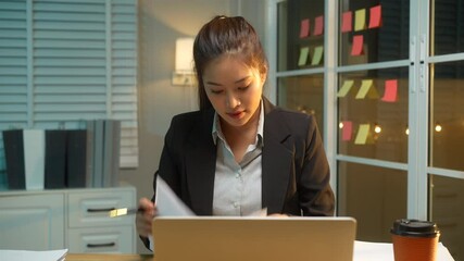 Business Asian woman in formal black suit thoughtfully reviews documents at her desk late at night, critical thinking, and commitment to meeting deadlines during late-night work hours.