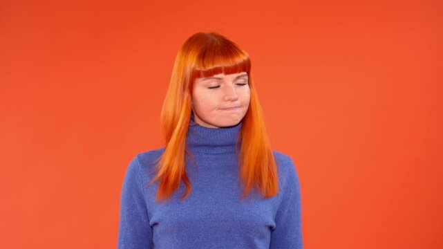 A young woman with vibrant orange hair and bangs, wearing a blue turtleneck sweater, stands against a bright orange background,showing disagreement and  shaking head side to side