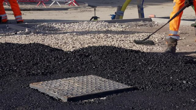 Road workers laying hot asphalt and prepare road surface around manhole cover at construction site