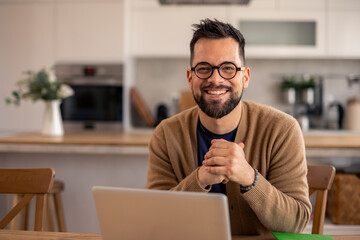 Smiling Man with Glasses Working on Laptop at Kitchen Table in Warm Bright Light