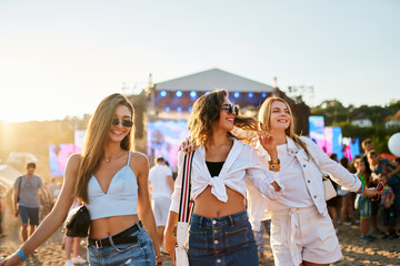 Three joyful young women stroll on sandy shore at sunset, friends laugh, dance at summer music...
