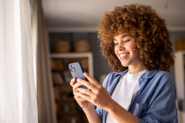 Smiling Woman with Curly Hair Holding Phone in a Bright and Cozy Living Room