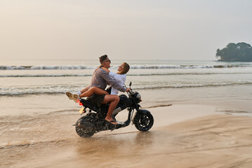 Obraz premium Couple rides motorbike on beach. Romantic getaway as woman hugs man on moving scooter. Love adventure by ocean. Fun and freedom on motorcycle journey together. Sand and waves around.