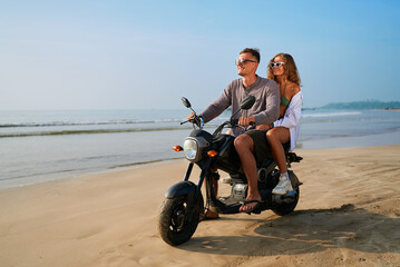 Obraz premium Couple enjoys beach ride on motorbike. Man drives motorcycle with woman on back. Ocean, sand, summer, sunglasses. Adventure travel, romantic journey, tropical holiday by sea.