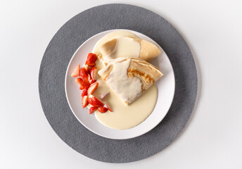 Plate with crepe pancakes, cut strawberries and vanilla sauce over everything on grey coaster on a white table top, flat lay