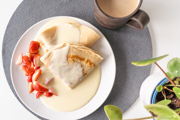 Crepe pancakes with cut strawberries and vanilla sauce with cafe latte on a grey coaster with a green plant on the side, flat lay
