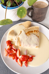 Side view of crepe pancakes with strawberries and vanilla sauce on a grey coaster with white coffee and a green plant on the side