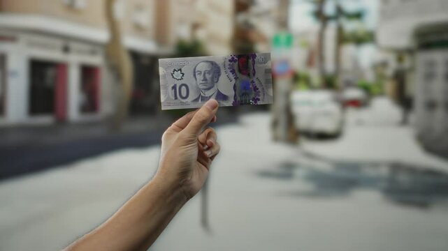 Hand holding canadian banknote outdoors on a blurred city street, capturing detail of a ten-dollar bill in a vibrant urban setting, emphasizing currency and male presence.