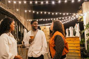 Three friends chatting and laughing together while enjoying drinks at a rooftop party under string...