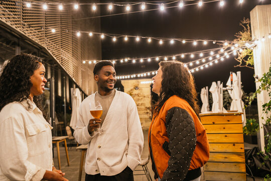 Three friends chatting and laughing together while enjoying drinks at a rooftop party under string lights