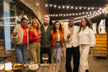 Multiethnic friends enjoying drinks and delicious food at a vibrant rooftop party, surrounded by twinkling string lights and city views