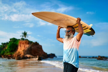 Active senior ethnic man carries surfboard on beach under blue sky. Elderly surfer enjoys ocean waves. Mature adventurer ready to surf, embrace spirit of surfing. Travel, explore coastal life.