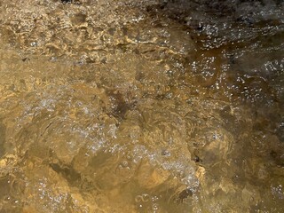 Close-up of water in a stream. Close-up and focused shot of the water surface in a small flowing stream. The reflection of light sparkles on the water surface flowing over natural rocks.