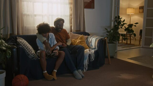 Full shot of modern African American father and his son laughing while enjoying video game together on cozy living room couch