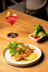 Pan-seared fish served on a plate with lettuce, cherry tomatoes, and sauce, placed on a wooden table with a red cocktail in the background for a refined seafood presentation.