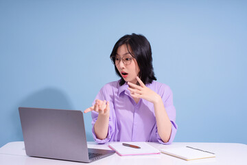 Young woman in purple shirt surprises during online meeting while sitting at the desk with laptop and notebooks in a light blue room