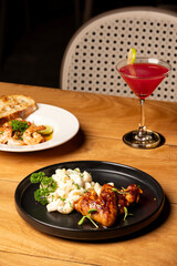 White cheese macaroni served with ginger roast chicken on a plate, with a red cocktail and prawns chimichurri in the background, all arranged on a table for a rich meal presentation.