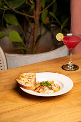 Prawns chimichurri served with bread on a plate, accompanied by a red cocktail on a table, with blurred green plants and a chair in the background for a cozy dining scene.