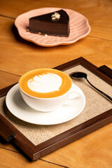 Obraz premium A cup of cappuccino with a spoon placed on a brown frame, with a slice of chocolate cake on a pink plate in the background, set on a brown restaurant table