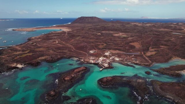4K aerial drone view of Puertito Isla de Lobos, Fuerteventura, featuring turquoise waters, rocky coastlines, volcanic terrain, and distant mountains that showcase coastal beauty and serene landscapes