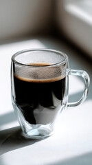 A transparent glass mug filled with dark, steaming coffee sits on a soft-lit surface, creating an inviting and cozy atmosphere