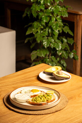 Avocado sandwich with toast and a half-fried egg omelette on a white plate, accompanied by a halved raw avocado on a small plate in the background, set on a table with blurred plants in a restaurant