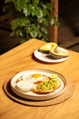 Avocado sandwich with toast and a half-fried egg omelette on a white plate, accompanied by a halved raw avocado on a small plate in the background, set on a table with blurred plants in a restaurant