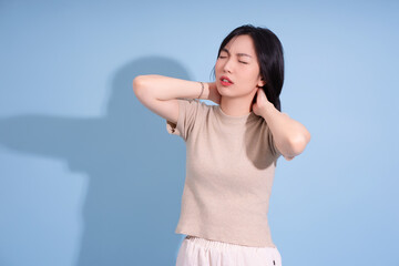 Young Asian woman experiencing neck pain while standing against a blue backdrop during daylight...