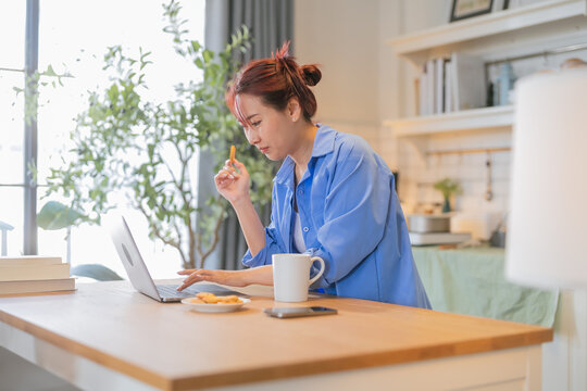 Young woman looking at laptop in kitchen  young Asian woman snacking on healthy rice crisps while working from home with laptop  working in kitchen with notebook 