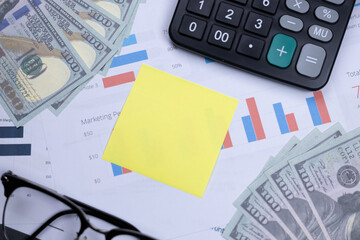 Fototapeta premium A yellow sticky note, cash, calculator, glasses, and financial charts on a desk, depicting business planning and financial analysis.
