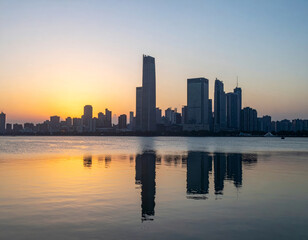 Fototapeta premium stunning city skyline at sunset, featuring tall buildings reflecting on calm water. warm hues of orange and blue create serene atmosphere, perfect for evening relaxation
