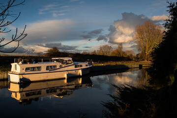Evening light over a cruiser moored on the River Lea, Enfield Lock. Dramatic clouds fill the sky and reflect on the calm water, creating a peaceful and atmospheric riverside scene.