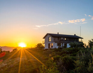 modern house situated on hill, bathed in warm glow of sunset, surrounded by lush greenery and distant mountains, creating serene and picturesque landscape
