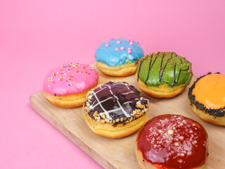 Colorful assorted donuts with vibrant icing and toppings on a wooden tray over a pink background