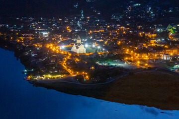 Night view of Mtskheta and Svetitskhoveli Cathedral from Jvari Monastery. The church is lit with warm light. Street and house lights are visible. Part of the Aragvi and Mtkvari (Kura) rivers.