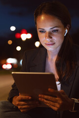 Urban businesswoman with wireless earphones using digital tablet at night in city street