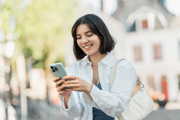 Mid-trip check-in with friends, multiracial Asian Latina Middle Eastern young woman with trendy short hair smiling and using phone in a European street