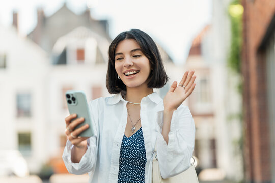 Booking a last-minute deal, short-haired traveler with Asian, Latina, and Middle Eastern roots smiles while using her phone in a lively European street