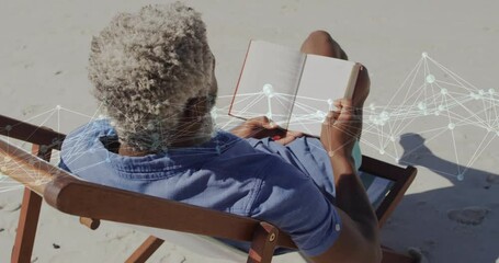 Senior man sitting reading hardcover book on sandy beach, showing technology network nodes overlay - Powered by Adobe