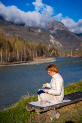 Fototapeta premium Woman playing a steel tongue drum in nature with a mountain river and Altai landscape in the background. Concept of harmony, mindfulness, and connection with nature