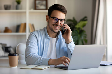 Smiling Caucasian Man with Glasses Talking on Phone and Using Laptop at Desk